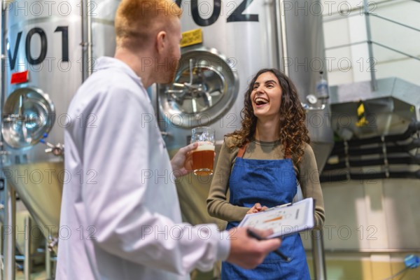Brewery worker and quality control specialist smiling, holding clipboard and a glass of beer, discussing brewing process in a modern industrial facility