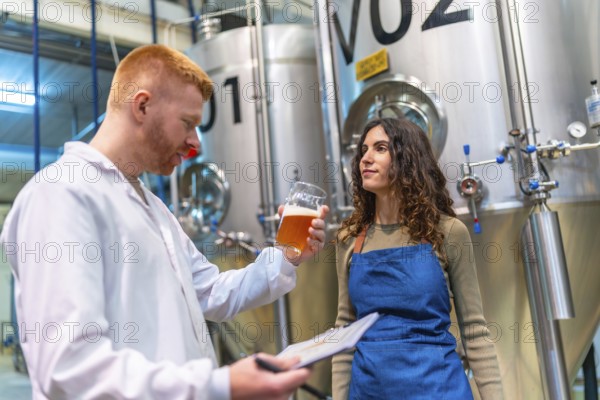 Brewery workers evaluating beer sample for quality control, holding clipboard and glass in brewing facility with large fermentation tanks, ensuring product excellence