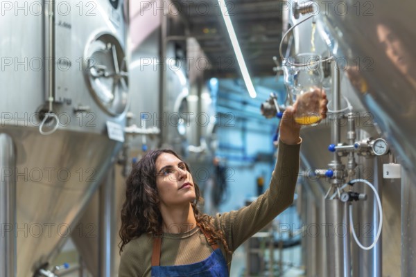 Woman brewer performing quality control, holding a glass sample of fresh beer, standing in a modern craft brewery facility with stainless steel fermentation tanks in the background