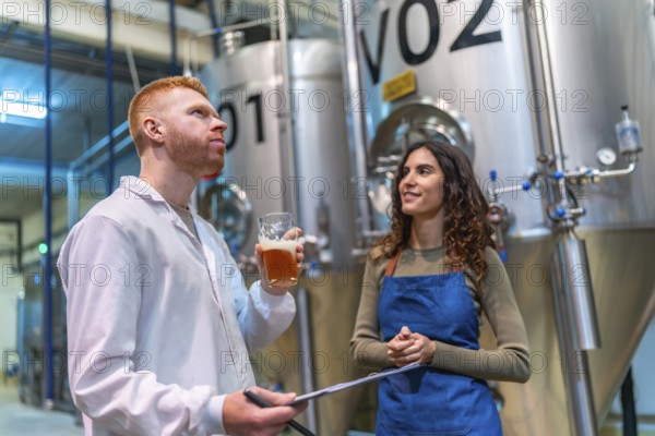 Two brewery professionals inspect and taste craft beer beside large fermentation tanks in a modern microbrewery, checking quality, recording results and collaborating on production