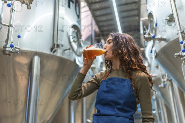Woman wearing an apron tasting a glass of freshly brewed beer for quality control, standing in a modern micro brewery with large fermentation tanks in the background
