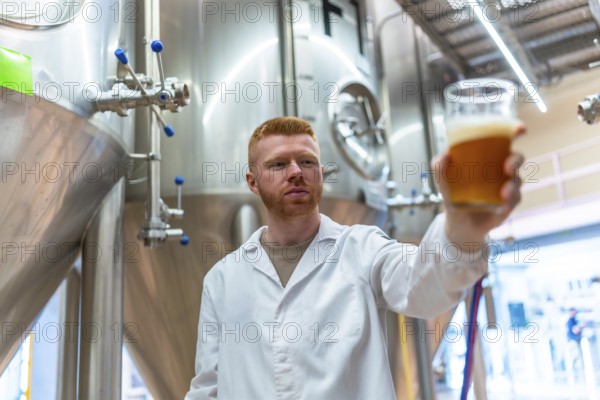 Brewery worker performing quality control by examining a glass of amber craft beer, ensuring production standards and evaluating the beverage for clarity, color, and foam in a factory setting