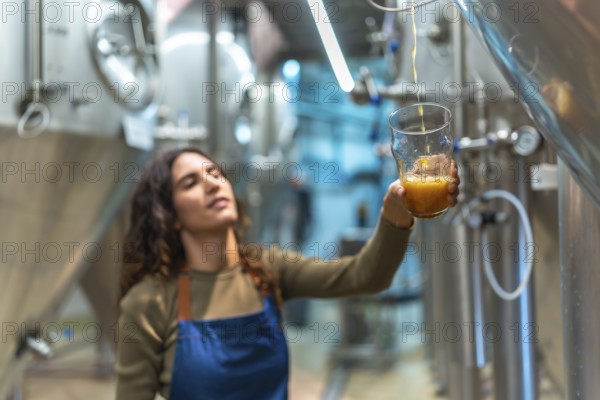 Female brewer wearing an apron, carefully pouring freshly brewed craft beer into a glass for a quality control taste test inside a modern brewery with stainless steel tanks