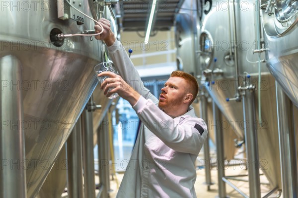 Brewery worker performing quality control, drawing a sample of beer for taste testing and analysis from a large stainless steel fermentation tank in a modern industrial setting