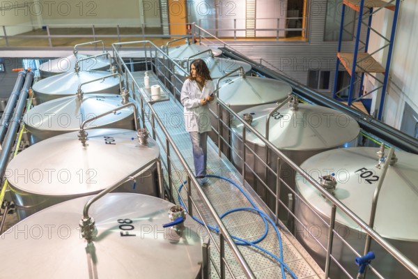 Woman scientist in a lab coat walking on a platform among large industrial fermentation tanks, inspecting operations and monitoring quality control in a modern beer brewery facility