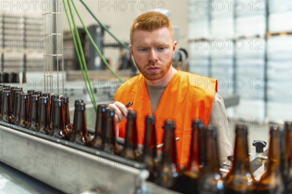 Man wearing an orange safety vest inspecting empty glass beer bottles moving along a conveyor belt in an industrial brewery facility, ensuring quality control