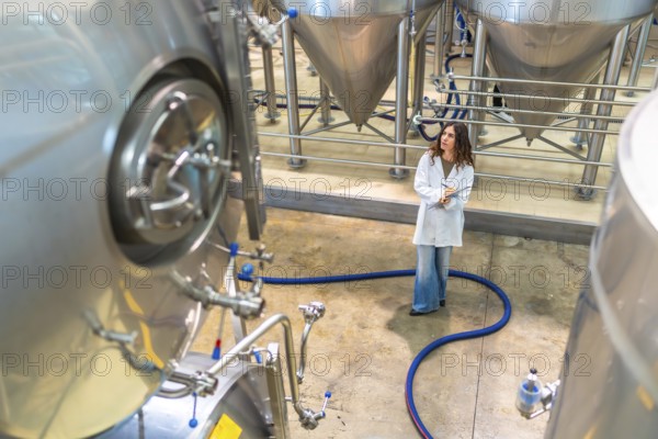 Female scientist wearing a lab coat and holding a clipboard, observing stainless steel fermentation tanks in a modern craft beer brewing facility, ensuring quality control and production