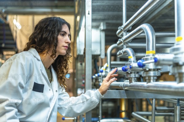 Young woman engineer wearing a lab coat and safety glasses. Meticulously adjusting valves on stainless steel pipes during a quality control inspection in a modern food or beverage production facility