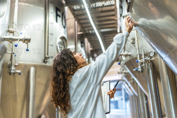 Young woman in a lab coat checking control panel on a stainless steel fermentation tank, performing quality control and inspection during the beer production process at a modern facility