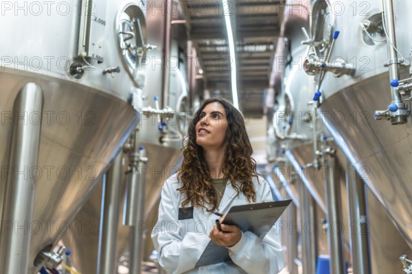 Woman scientist in a white lab coat with clipboard conducting quality control inspection in a modern craft beer production facility among stainless steel fermentation tanks and equipment