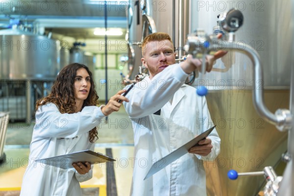 Chemical engineers and a quality control technician are checking stainless steel fermentation tanks and valves in a modern beer factory, ensuring production standards and process efficiency