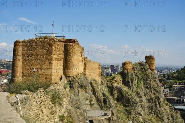 A weathered stone fort stands on a rocky hill, crowned with a cross. Below, a vibrant city stretches out under a clear blue sky, showcasing a mix of modern buildings and nature