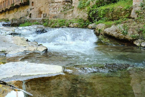 Gentle water flows over smooth stones revealing a tranquil scene. Surrounded by ancient walls and lush greenery, the area invites peaceful reflections. Nature thrives in this picturesque setting