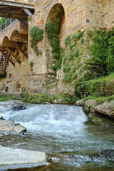 Clear water streaming over smooth stones next to an ancient stone wall adorned with lush plants. Sunlight illuminates the peaceful landscape, enhancing its natural beauty