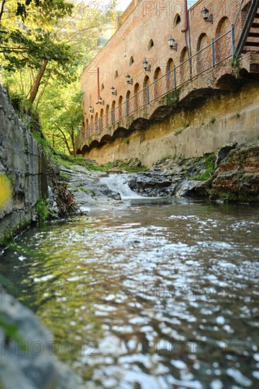 A clear stream flows calmly past old brick structures surrounded by lush greenery. Sunlight sparkles on the water's surface, creating a serene atmosphere in this peaceful outdoor scene