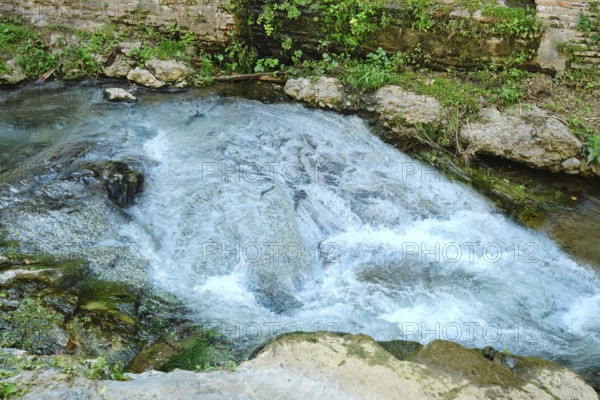 A small river flows over smooth stones, creating gentle ripples and splashes. Surrounded by greenery and natural rocks, this peaceful scene captures the beauty of nature on a bright day