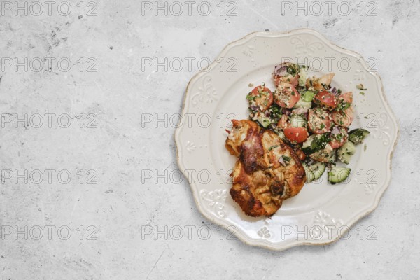 A plate showcases succulent roasted chicken pieces beside a fresh salad made with tomatoes, cucumbers, and herbs. The light-colored dish rests on a textured gray surface, adding elegance