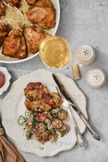 Grilled chicken pieces are plated next to a vibrant salad made of cucumbers and tomatoes. A glass of white wine completes the meal. The table is set for a lovely dining experience