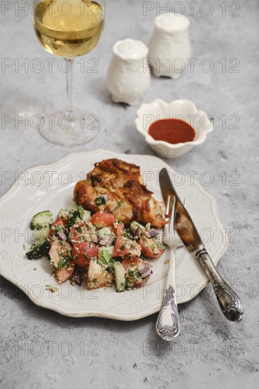 A well-prepared meal features grilled chicken, fresh salad with tomatoes and cucumbers, and a glass of white wine. The setting includes small condiment containers and a vintage plate