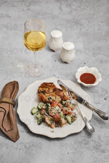 A beautifully arranged meal includes a golden glass of wine, a serving of baked meat, fresh salad with herbs, and a small bowl of sauce. A cloth napkin adds a touch of elegance to the table setting