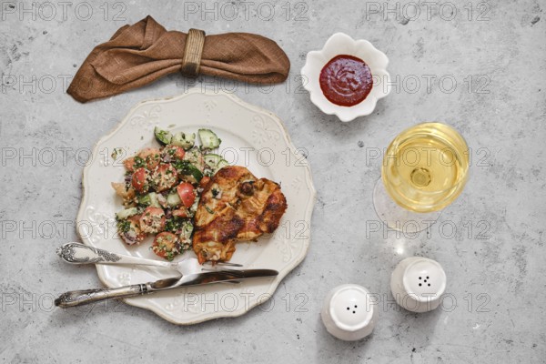 A beautifully arranged meal features grilled chicken with a side of vibrant salad made of tomatoes and cucumbers. A glass of white wine complements the dish, alongside a small bowl of sauce