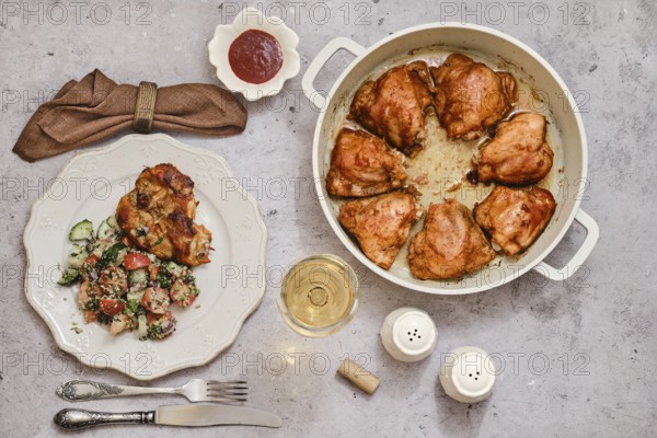 A well-cooked dish of roasted chicken thighs in a large pot, accompanied by a vibrant salad of cucumber and tomatoes on a plate, alongside a glass of white wine, ready for dinner