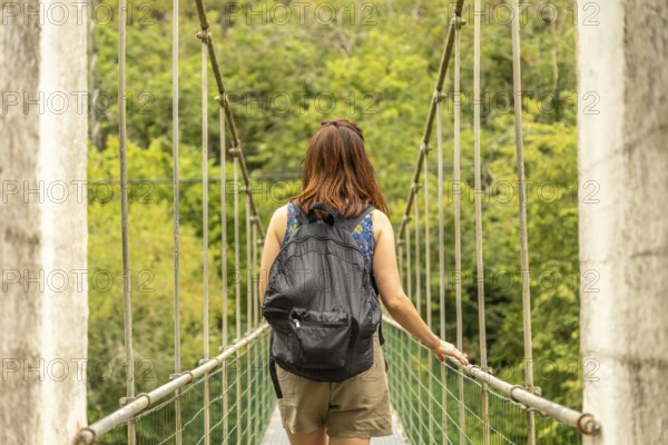 Female tourist with backpack walking on a hanging bridge over the sella river, surrounded by lush green forest in asturias, spain, enjoying a summer adventure