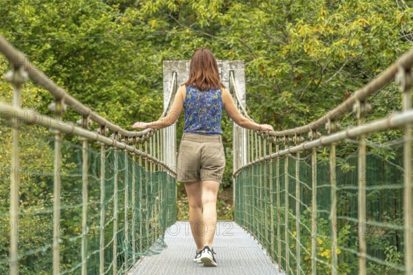 Female tourist enjoying a peaceful walk across a metallic hanging bridge, surrounded by lush green vegetation, on the route from the dobra river to the olla de san vicente in asturias, spain