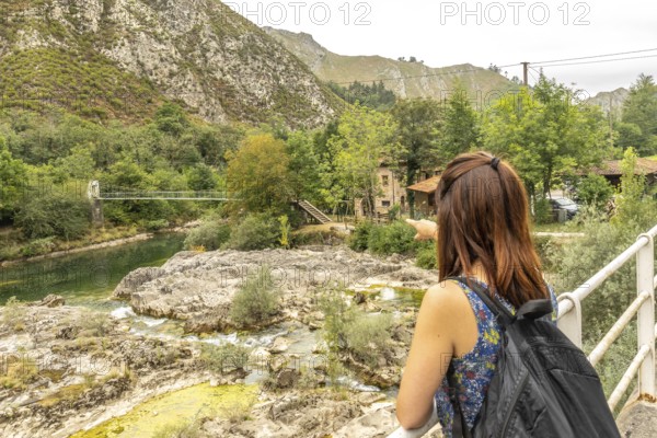Tourist with a backpack pointing at the olla de san vicente, a natural pool on the sella river, surrounded by mountains and vegetation in sunny asturias, spain