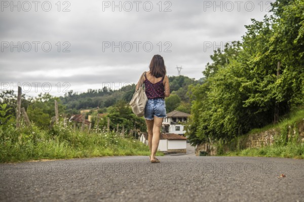 Young woman tourist walking on a road approaching benia de onis village in asturias, spain, during a cloudy summer day, carrying a tote bag