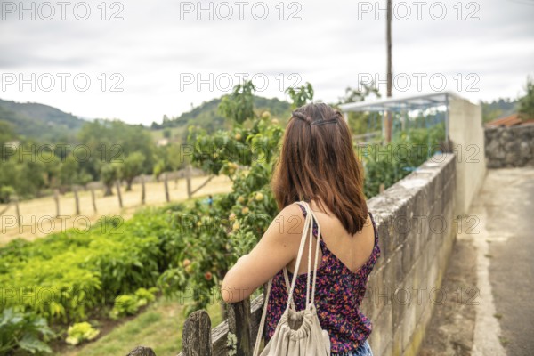 Young woman tourist with backpack is enjoying the view of a peach orchard and a vegetable garden in the picturesque village of benia de onis, asturias, spain, during a summer day
