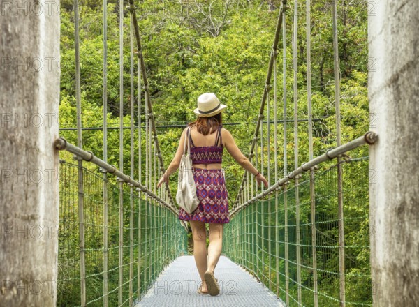Young woman tourist with a hat and bag walking on a hanging bridge over the dobra river, enjoying a scenic summer adventure in asturias, spain