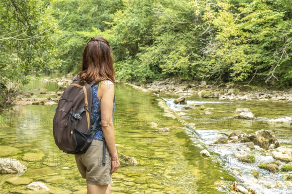 Female tourist with a backpack admiring the dobra river along the hiking route to olla de san vicente, surrounded by lush vegetation on a sunny summer day in asturias, spain