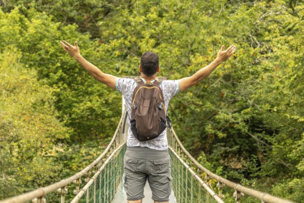 Backpacker opening his arms while enjoying the view from a hanging bridge over the sella river, surrounded by the lush vegetation of asturias, spain, during a sunny summer day