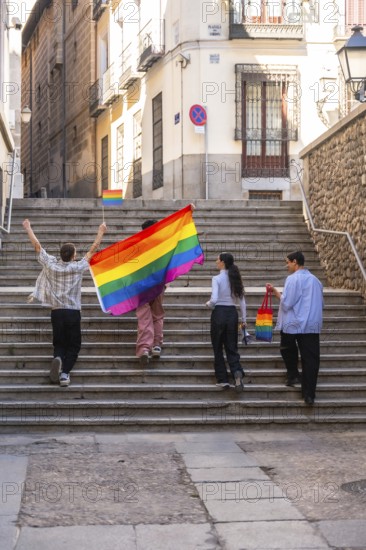Group of diverse friends celebrating pride, walking up old stairs in a city, carrying a large rainbow flag and displaying a rainbow themed bag, expressing joy