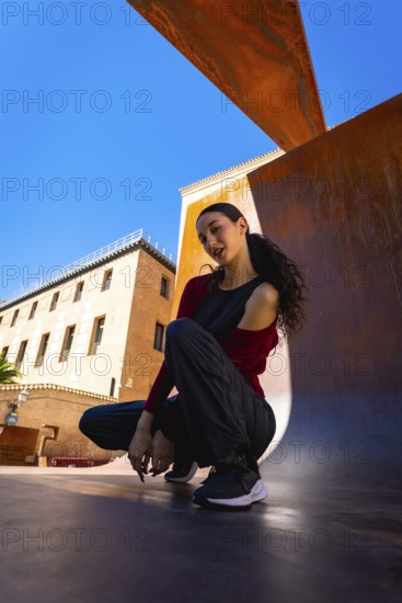 Young woman squatting on an urban surface, posing casually against a modern rust colored architectural element with old city buildings and a clear blue sky in the background