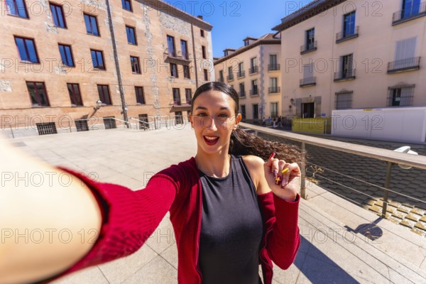 Young woman smiling and holding a smartphone, capturing a selfie portrait in an urban setting surrounded by historic european buildings on a sunny day during her city trip