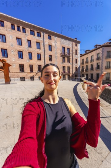 Young woman with dark hair posing for a selfie in a city square, holding her hand up, with old european buildings and a clear blue sky in the background