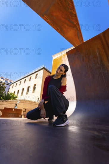 Young woman posing in a modern city environment, featuring rust colored metal structures and historical buildings under a clear blue sky on a bright day