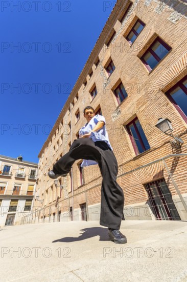 Young man doing a martial arts kick pose with focus and determination, dressed in casual loose clothing on a sunny day in an urban street with a brick building in the background