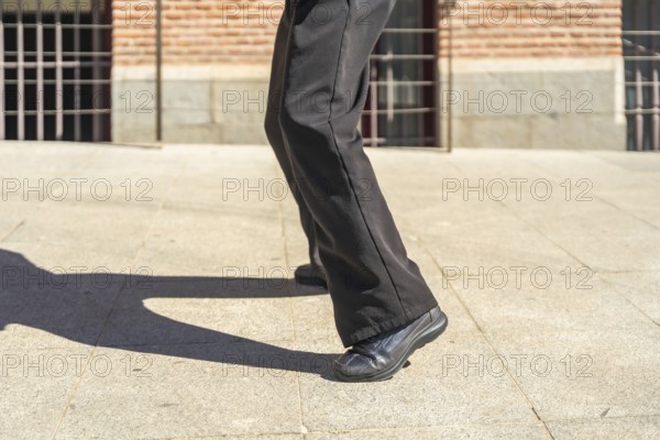 Person's legs moving while dancing outdoors, casting a long shadow on the light colored pavement of an urban street with a brick building in the background