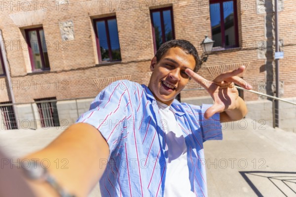 Young man smiling and making a playful hand gesture while taking a selfie in sunny urban streets, enjoying city architecture and capturing travel vibes for social media