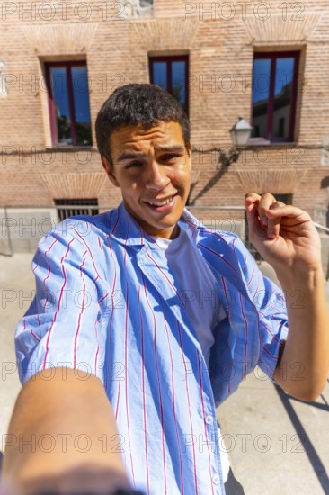Young man smiling directly at the camera while taking a selfie outdoors on a sunny day, wearing a striped shirt and standing in front of an old brick building