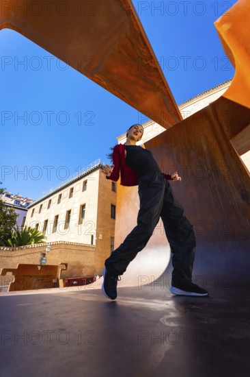 Woman performing a dynamic dance move, expressing freedom and urban culture against a vibrant blue sky and a modern rusty metal art sculpture in a city environment