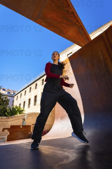 Young woman moving energetically, practicing dance in an urban setting, with modern corten steel architecture and a clear blue sky creating a dynamic scene