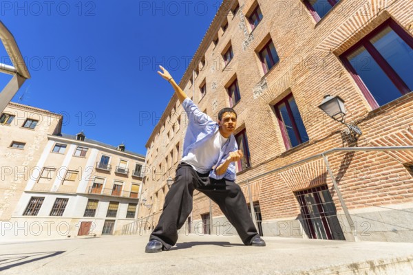 Young man actively posing or dancing in a dynamic stance on a sunny urban street, expressing freedom and joy against a backdrop of traditional brick architecture and a clear blue sky