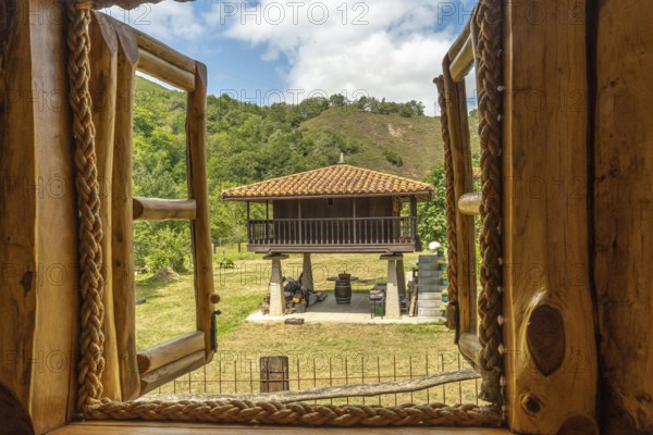 Open wooden window with rope hinges framing a scenic view of a traditional elevated horreo granary in the serene countryside of asturias, spain