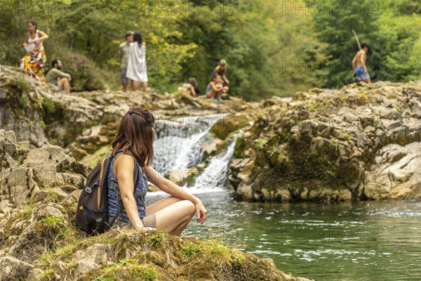 Young female hiker resting on rocks near a small waterfall on the dobra river, with other hikers enjoying the natural pool in the background, on a sunny day in asturias, spain