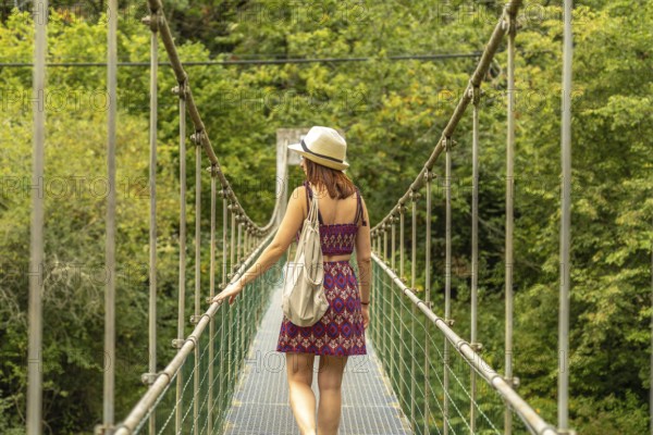 Female tourist with hat and backpack walking on a metal suspension bridge, enjoying the scenic beauty of a lush green forest, experiencing the thrill of adventure and exploration in nature