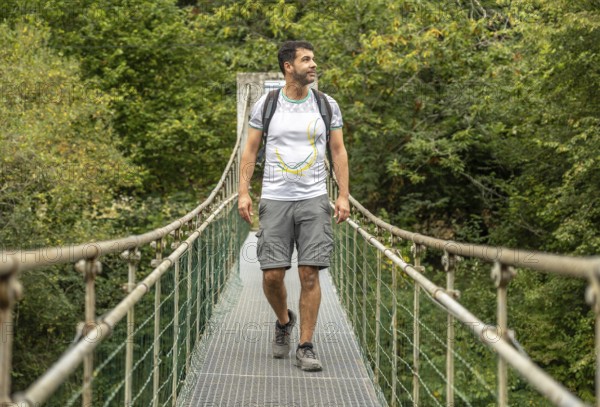 Male hiker with backpack enjoying trekking in the forest, walking on a metallic hanging bridge over a river, surrounded by lush green vegetation